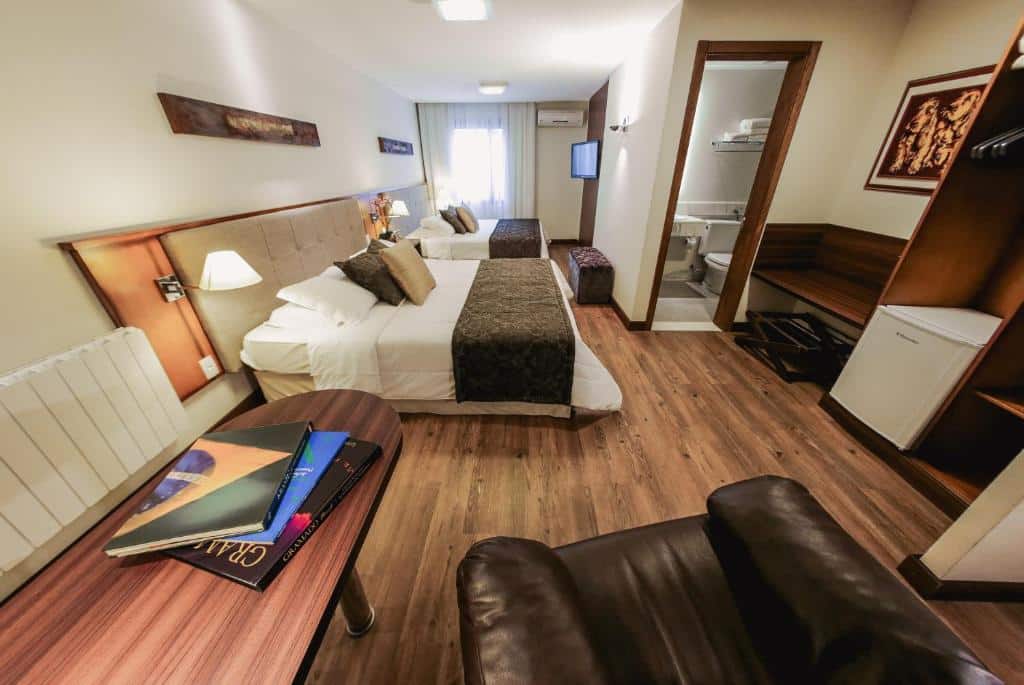 quarto do bavaria sport hotel com duas camas de casal, lado lado, uma televisão de tela plana, uma poltrona de courino marrom e uma porta com acesso ao banheiro no lado direito da imagem.