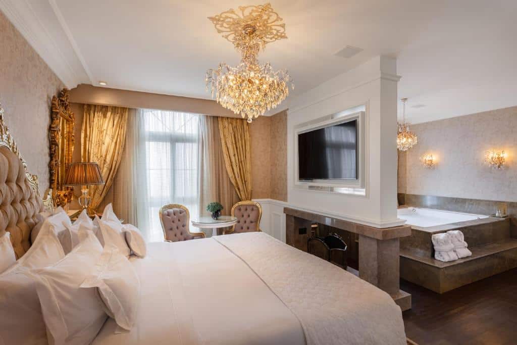quarto do hotel colline de france para representar onde ficar em Gramado. A suíte é decorada com detalhes dourados nas cortinas, na cabeceira da cama de casal, nas cadeiras e nas arandelas do teto. Há ainda uma banheira de hidromassagem e uma lareira que divide o quarto