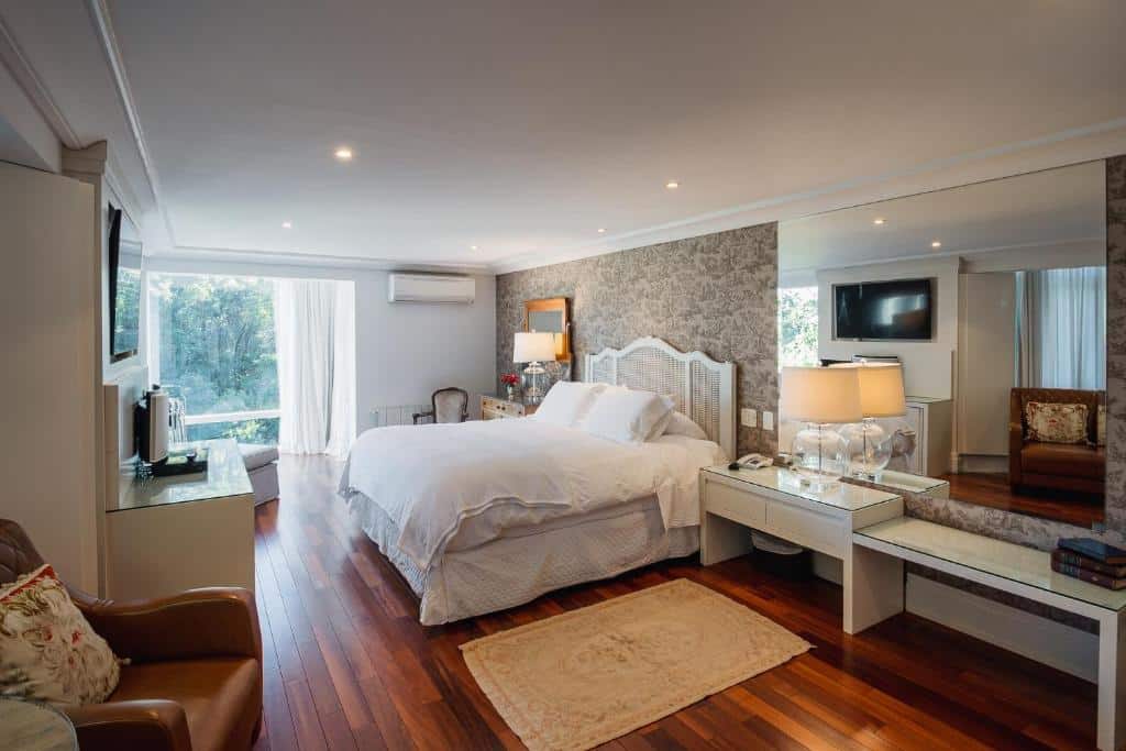 quarto do hotel estalagem st hubertus com uma cama de casal no centro do espaço, uma cômoda branca pequena ao lado da cama, um espelho retangular e grande em cima da comoda cobrindo boa parte da parede, e uma janela ampla do teto ao chão no canto esquerdo com vista para as árvores