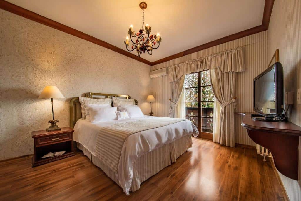 suíte do hotel casa da montanha com uma cama de casal, um lustre delicado e trabalhado no teto, uma porta dupla com acesso à varanda particular e uma televisão no lado direito da imagem.