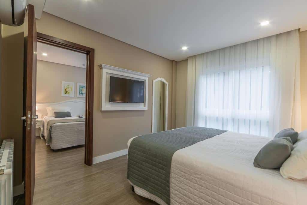 quatro duplo do hotel monte felice bosque mostrando uma cama de casal à direita da imagem com uma televisão pendurada na parede em frente a cama, ao lado da televisão há uma porta aberta onde é possível ver outro quarto com uma cama de solteiro.