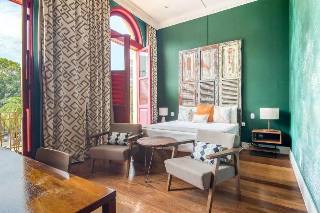 Um quarto do Selina Lapa com paredes verdes, uma cama de casal, duas poltronas, uma pequena mesa e grandes portas com janelas que dão para uma varanda. Essa é a uma das opções de onde ficar no Rio de Janeiro.