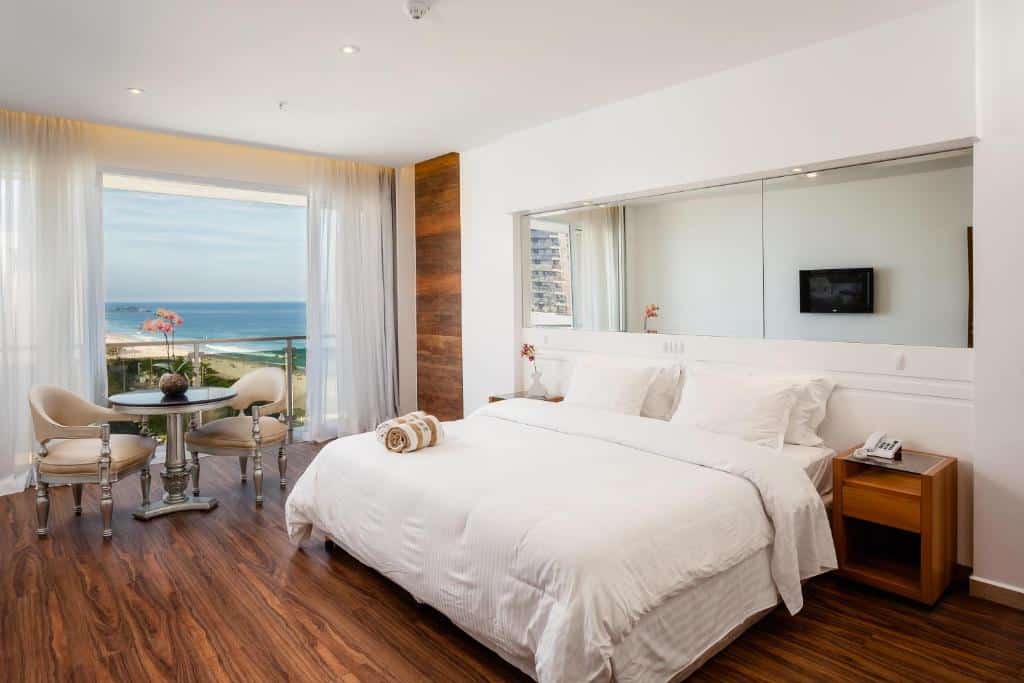 Um quarto do Praia Ipanema Hotel com uma cama grande, mesa de jantar, janela com vista para o mar, piso de madeira e TV na parede. Essa é a uma das opções de onde ficar no Rio de Janeiro.