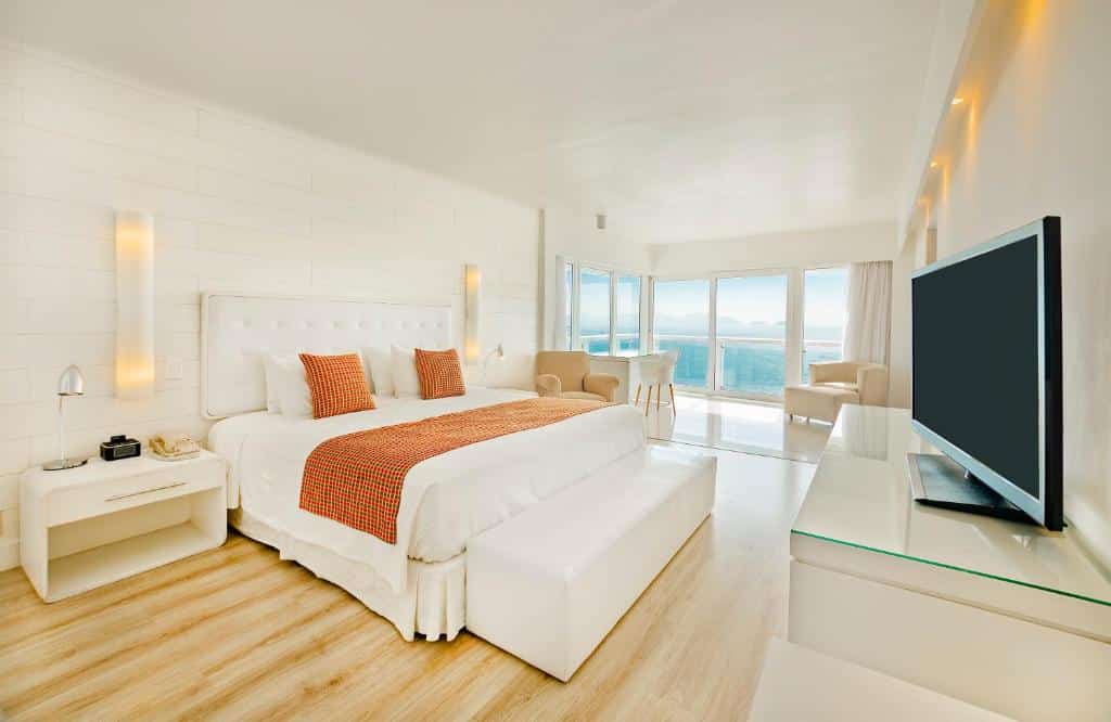 Quarto do Rio Othon Palace bem iluminado com uma cama grande, travesseiros laranja, TV, cadeiras e janelas grandes com vista para o oceano. Essa é a uma das opções de onde ficar no Rio de Janeiro.