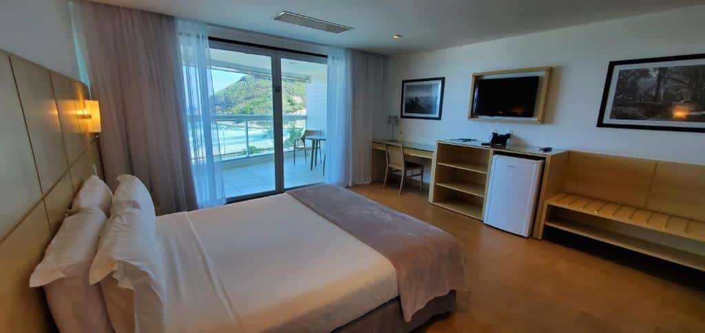 Quarto do Atlântico Sul Hotel com cama de casal, escrivaninha, TV, frigobar e varanda com vista para a praia e as colinas. Essa é a uma das opções de onde ficar no Rio de Janeiro.