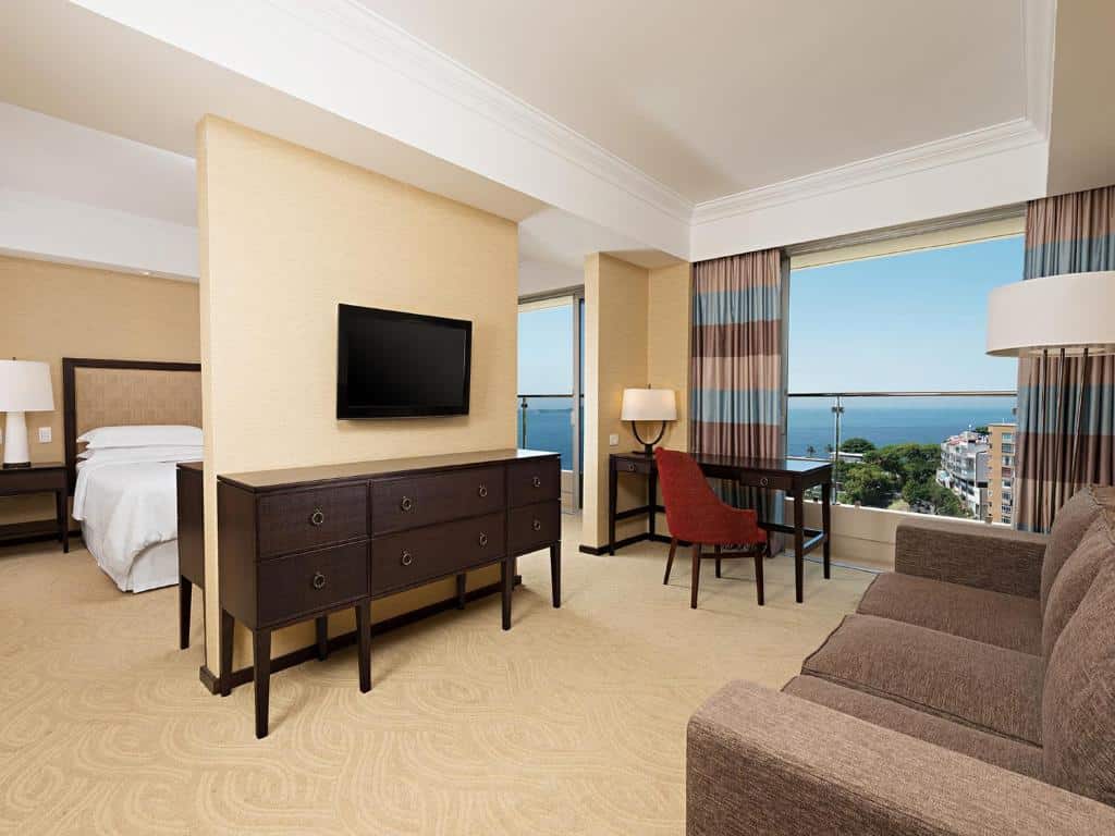 Suíte do Sheraton Grand Rio Hotel & Resort com cama, sofá, TV, escrivaninha, janelas grandes e vista para os prédios e o mar. Essa é a uma das opções de onde ficar no Rio de Janeiro.