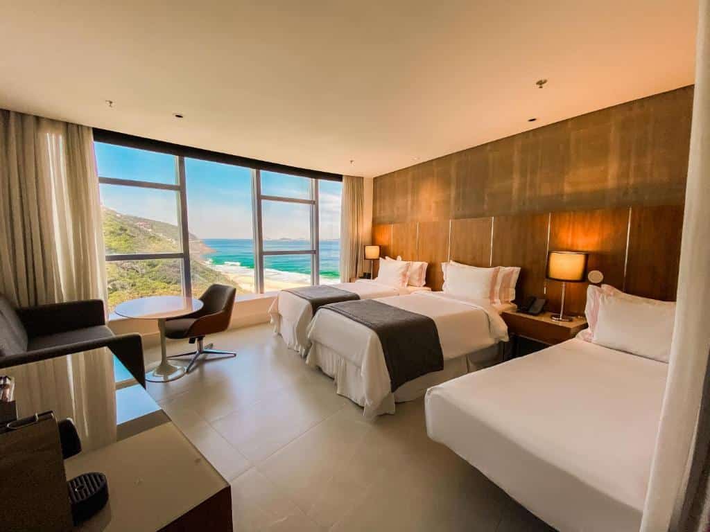 Quarto do Hotel Nacional com três camas, janelas grandes, vista para o mar e para a montanha, pequena mesa redonda, cadeiras e luminárias. Essa é a uma das opções de onde ficar no Rio de Janeiro.