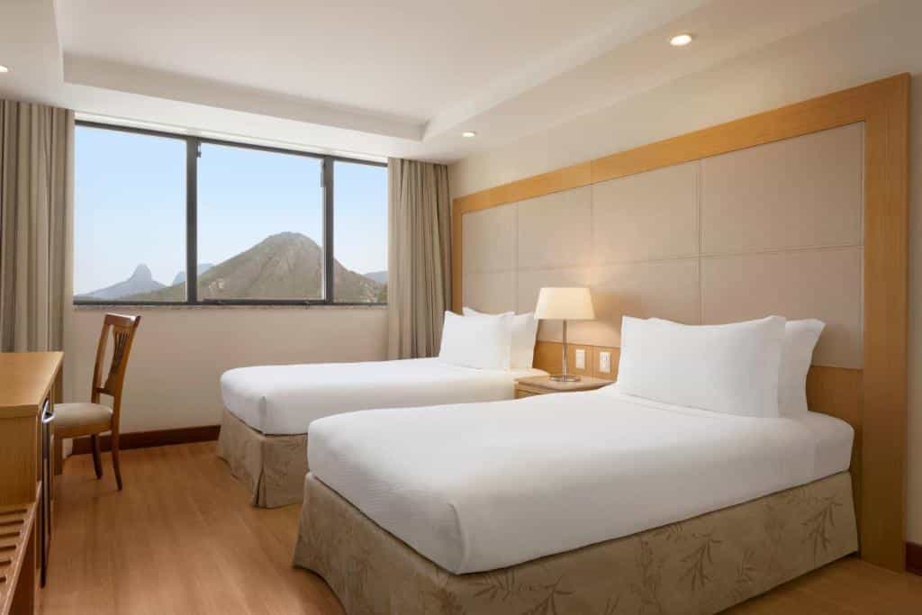 Quarto do Hilton Copacabana com duas camas de solteiro bem arrumadas, uma escrivaninha e cadeira, um abajur e uma grande janela com vista para a montanha. Essa é a uma das opções de onde ficar no Rio de Janeiro.