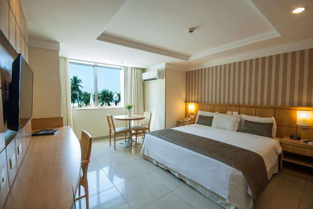 Quarto do Hotel Astoria Palace com uma cama grande, escrivaninha, mesa e cadeiras, luminárias, TV e uma janela com palmeiras do lado de fora. Essa é a uma das opções de onde ficar no Rio de Janeiro.