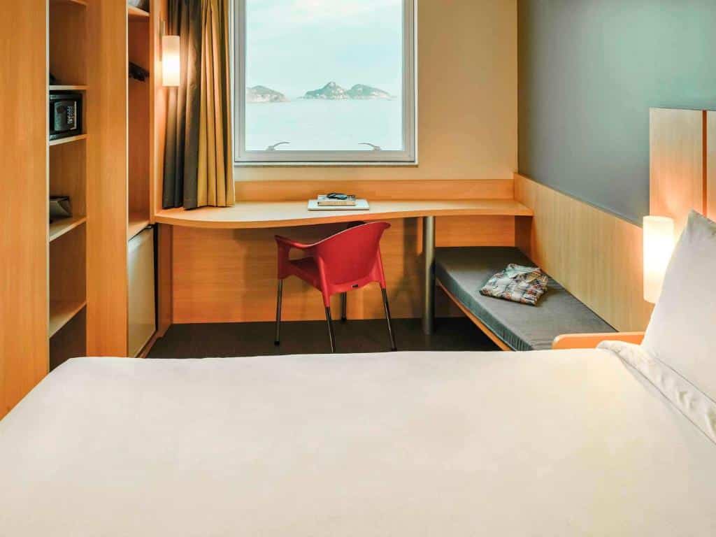 Um pequeno quarto do ibis Barra da Tijuca com uma cama, escrivaninha, cadeira vermelha, banco, livros e uma janela com vista para ilhas distantes. Essa é a uma das opções de onde ficar no Rio de Janeiro.