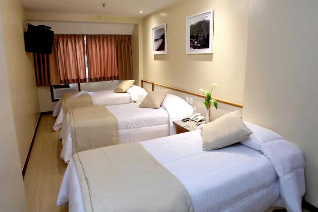 Quarto do Mengo Palace Hotel com três camas de solteiro, roupa de cama bege, duas fotos emolduradas na parede e cortinas cobrindo a janela. Essa é a uma das opções de onde ficar no Rio de Janeiro.