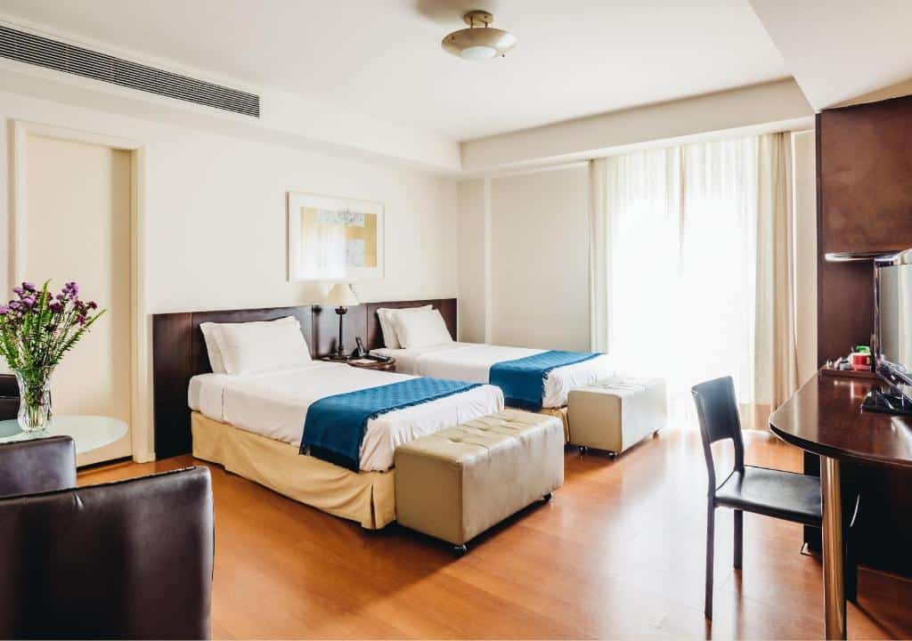 Quarto do Own Ipanema Visconti com duas camas de solteiro, corredores azuis, uma TV, escrivaninha, cadeira marrom e luz do sol entrando pelas cortinas transparentes. Essa é a uma das opções de onde ficar no Rio de Janeiro.