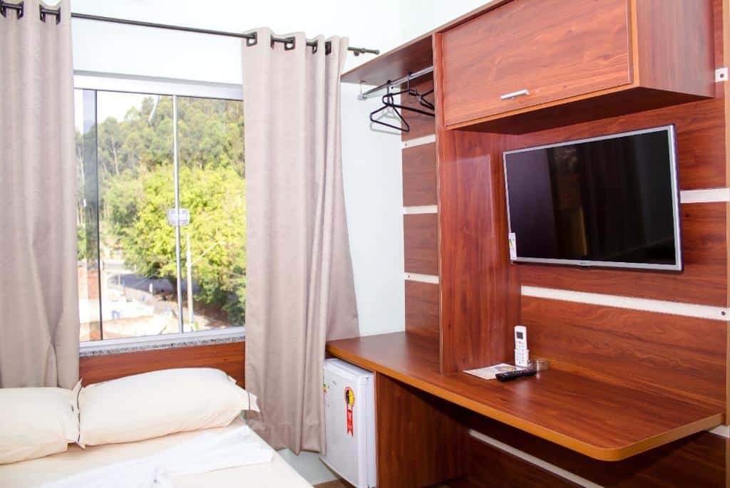 Quarto pequeno da Pousada Monte Oliveira com uma cama, TV na parede, frigobar, escrivaninha e uma janela com cortinas com vista para as árvores do lado de fora.