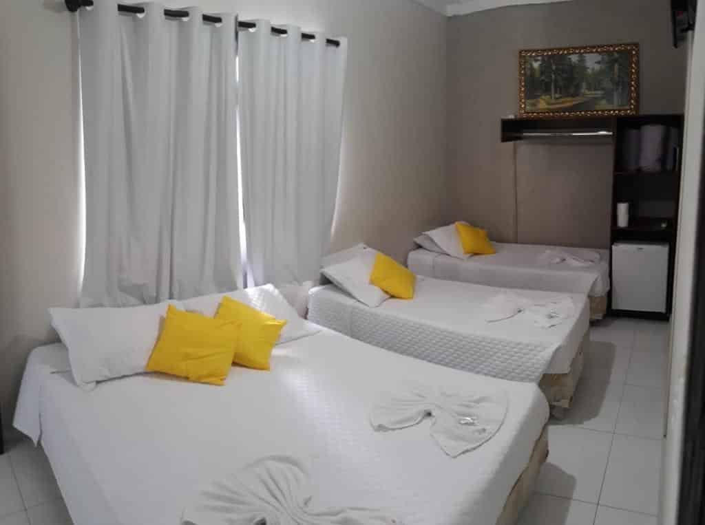 Quarto da Pousada Aconchego com três camas de solteiro, lençóis brancos, travesseiros amarelos e um quadro na parede.