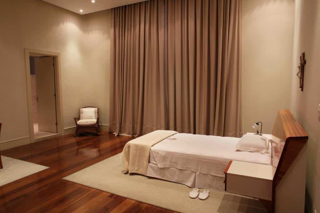 Quarto minimalista da Pousada do Bom Jesus com uma cama de solteiro, piso de madeira, cortinas bege, uma cadeira e um par de chinelos brancos no tapete.