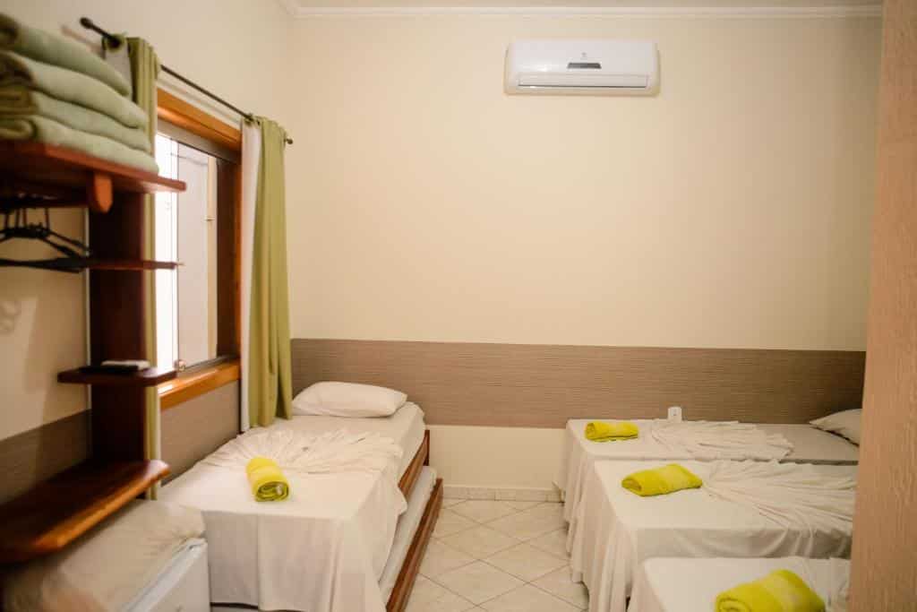 Três camas de solteiro na Pousada Santa Cruz com lençóis brancos e toalhas amarelas em um quarto pequeno e simples com ar-condicionado e janela.