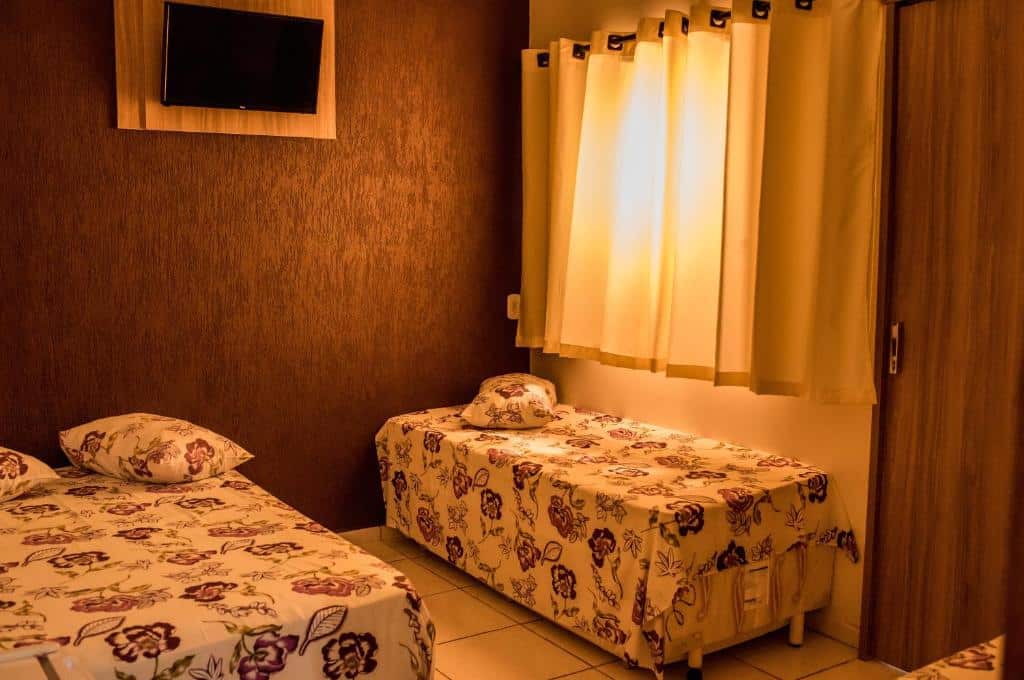 Duas camas de solteiro com roupa de cama floral, uma TV na parede, cortinas fechadas e paredes marrons em um quarto simples da Pousada Rabake.
