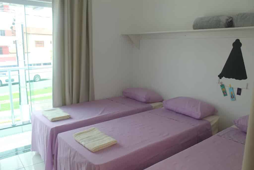 Um quarto da Pousada Orquideas de Maria com duas camas de solteiro com roupa de cama roxa, uma prateleira com toalhas e uma janela com cortinas bege.