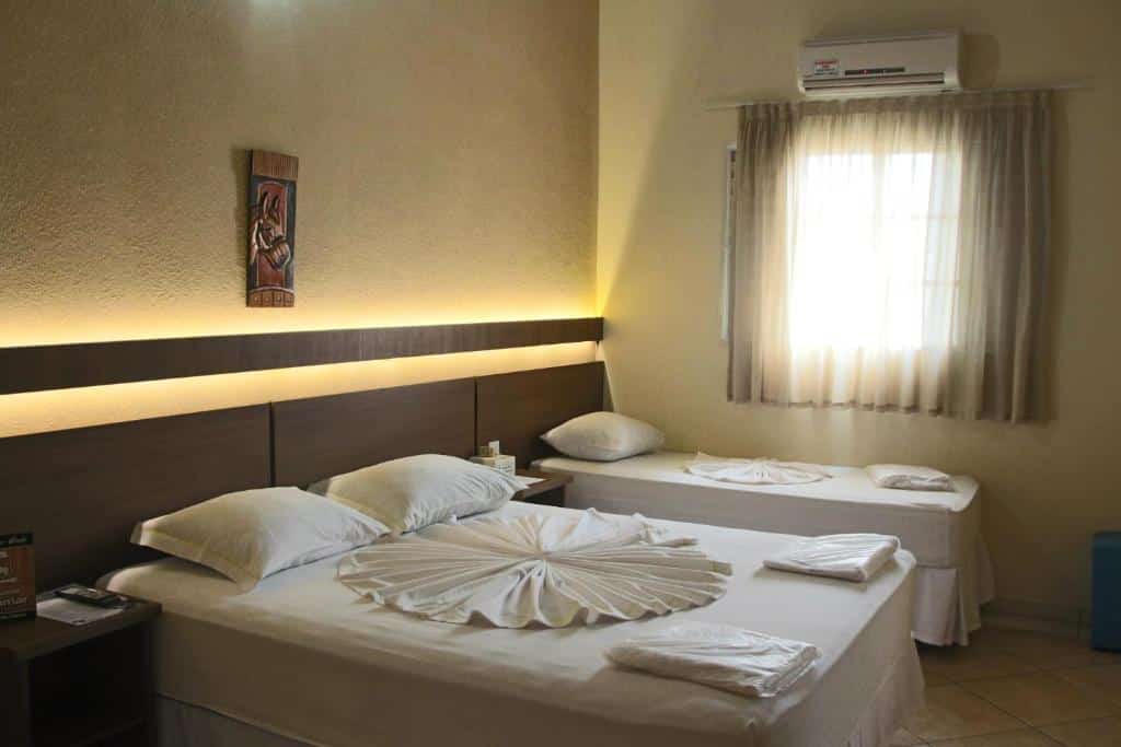 Um quarto da Pousada Jovimar com uma cama de casal e uma cama de solteiro, ambas com lençóis brancos, perto de uma janela com cortinas transparentes.