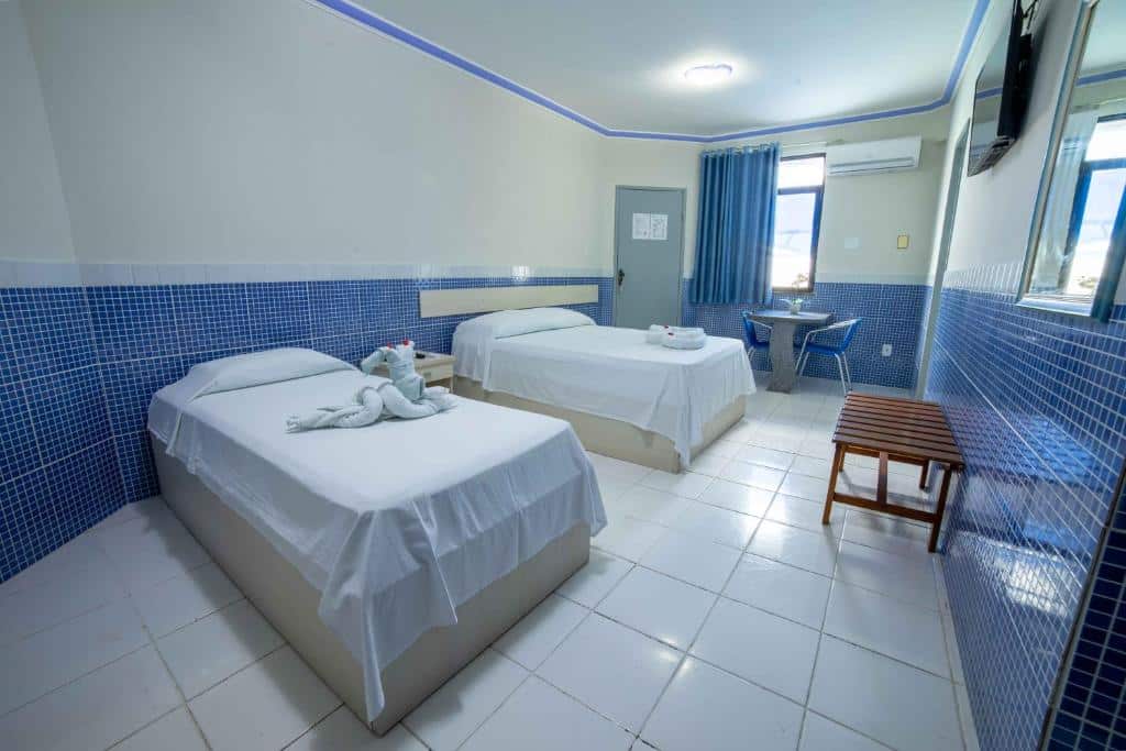 Quarto do Hotel Pousada do Farol com duas camas, mesinha com cadeiras, locai para apoiar a mala, ar-condicionado e TV