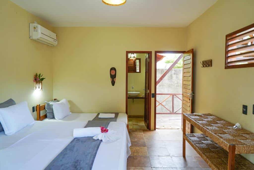 Um quarto da Pousada Rota dos Ventos simples com uma cama de casal, móveis de vime, uma porta aberta para um pátio e paredes de cores neutras. Representa o post de pousadas em Barra Grande no Piauí.