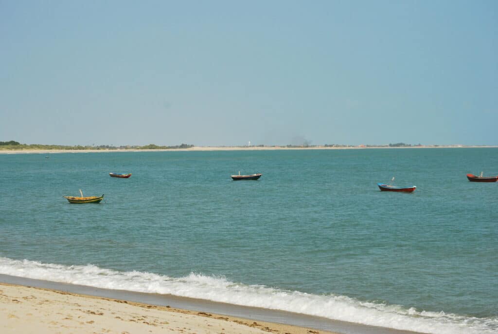 Vários barcos pequenos flutuam em águas azuis calmas perto de a praia de Barra Grande no Piauí sob um céu claro.