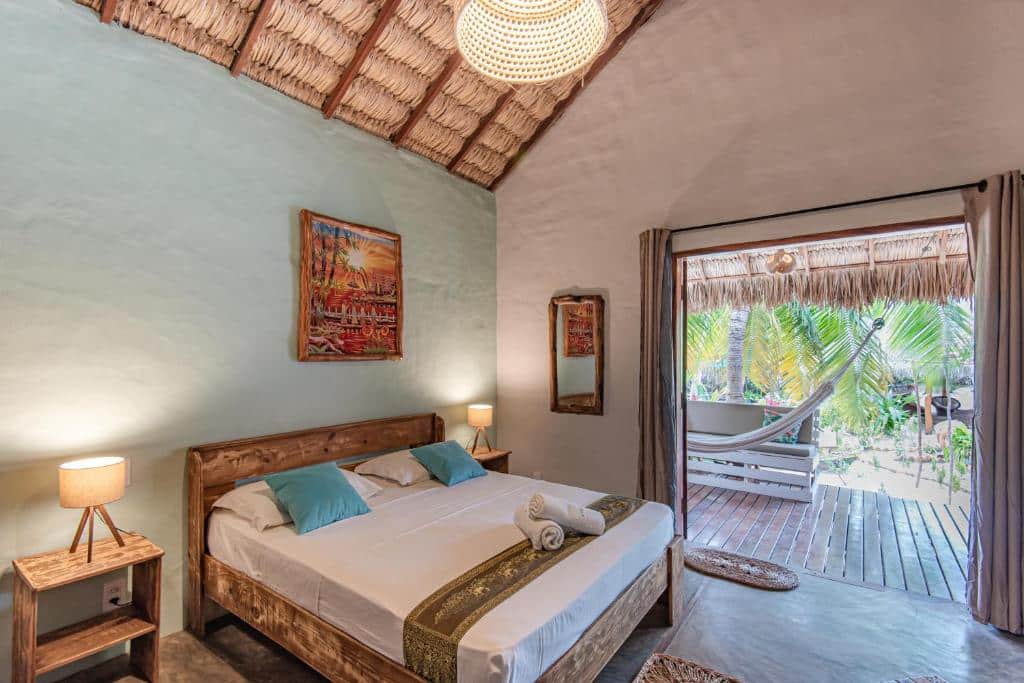 Um quarto rústico da Pousada Villa Zen com uma cama de casal, móveis de madeira, luminárias, obras de arte e uma porta aberta que dá para um pátio com rede. Representa o post de pousadas em Barra Grande no Piauí.