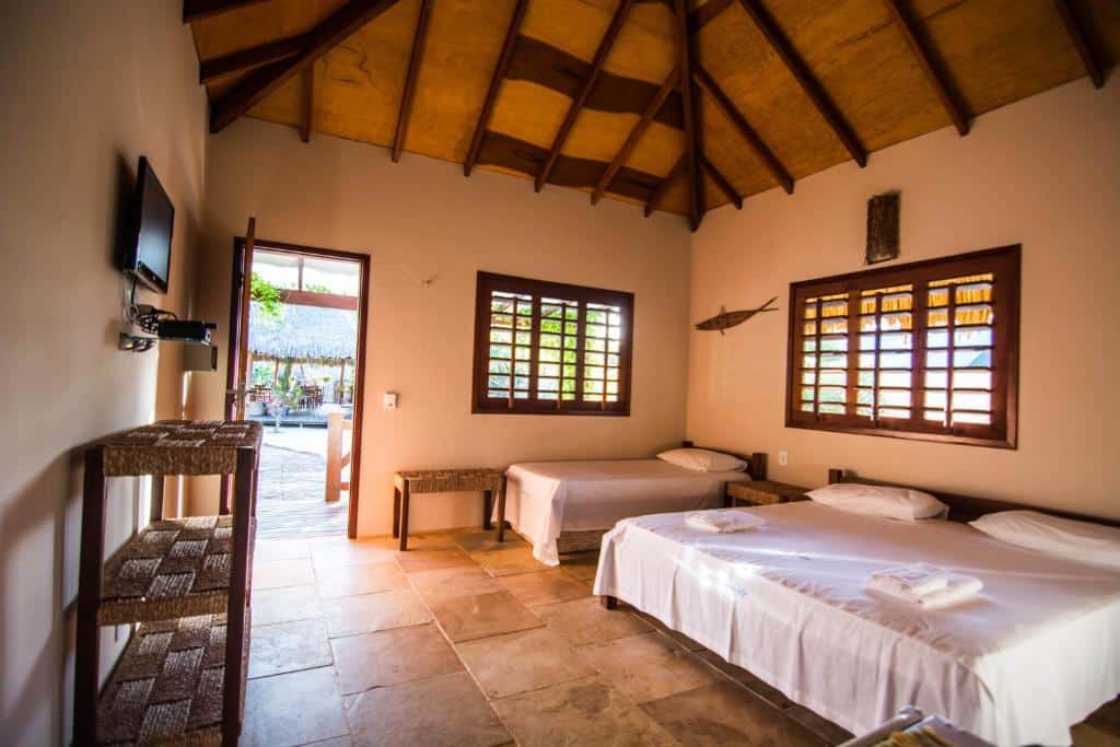 Um quarto simples da Pousada Vento do Kite com duas camas, uma TV na parede, janelas com persianas de madeira e uma porta que dá para uma área externa. Representa o post de pousadas em Barra Grande no Piauí.