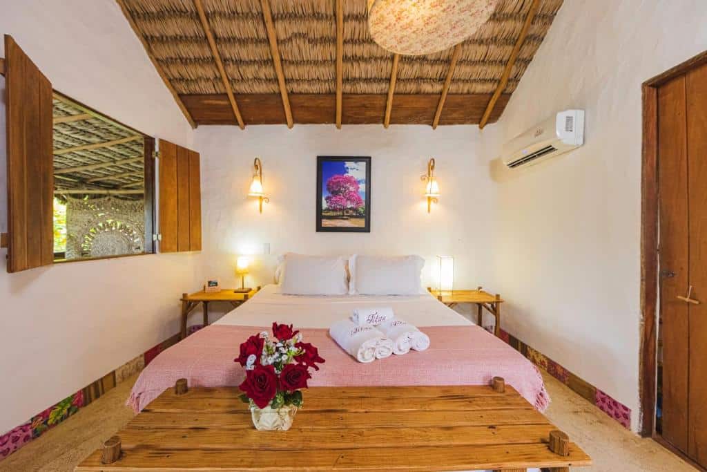 Um quarto da Pousada Titas com uma colcha rosa, toalhas, flores em um banco de madeira, duas lâmpadas e uma foto emoldurada acima da cama. Representa o post de pousadas em Barra Grande no Piauí.