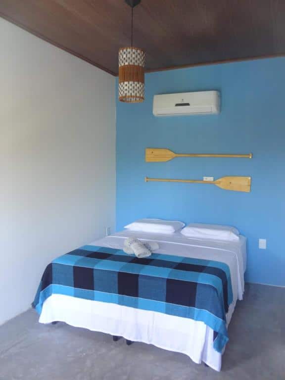 Quarto da Pousada Angels com uma cama com um cobertor xadrez azul e preto, dois travesseiros e remos montados em uma parede azul acima da cabeceira. Representa o post de pousadas em Barra Grande no Piauí.