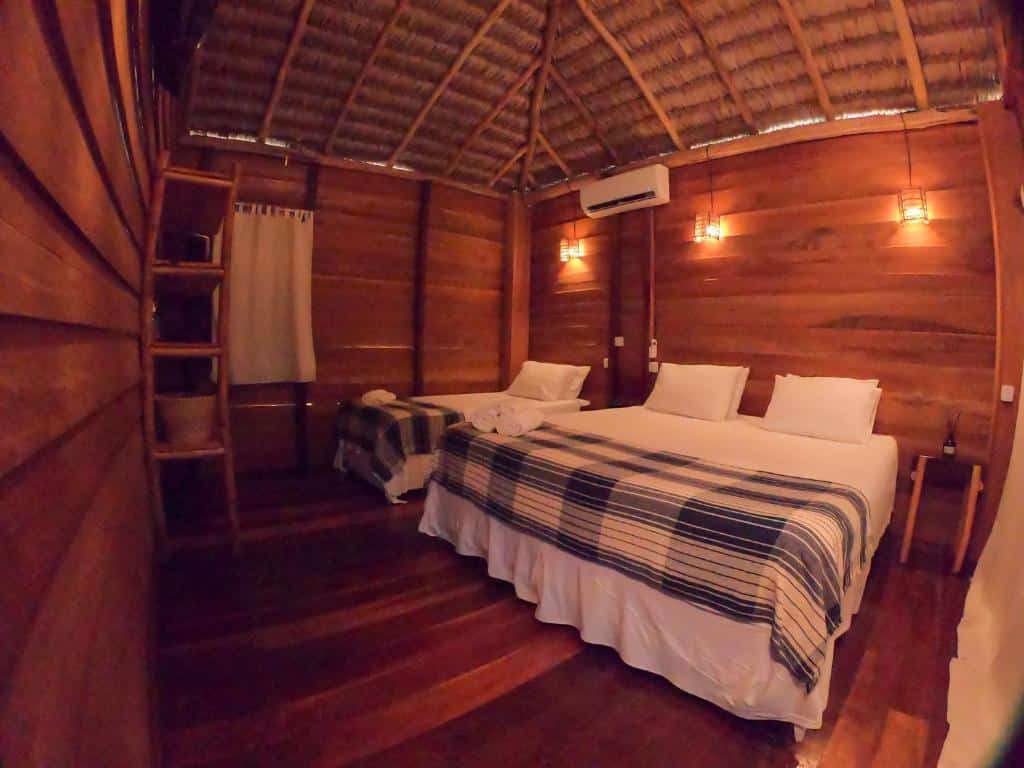Quarto da Na Praia Pousada com painéis de madeira, duas camas, roupa de cama listrada, uma pequena janela, luzes de parede e uma escada com prateleira de madeira. Representa o post de pousadas em Barra Grande no Piauí.