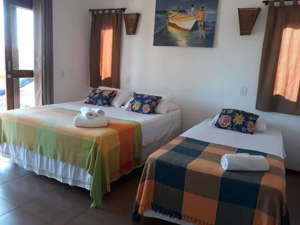 Um quarto da Pousada Lá em Casa com uma cama de casal e uma cama de solteiro, ambas com cobertores e toalhas coloridas, e um quadro acima das camas. Representa o post de pousadas em Barra Grande no Piauí.