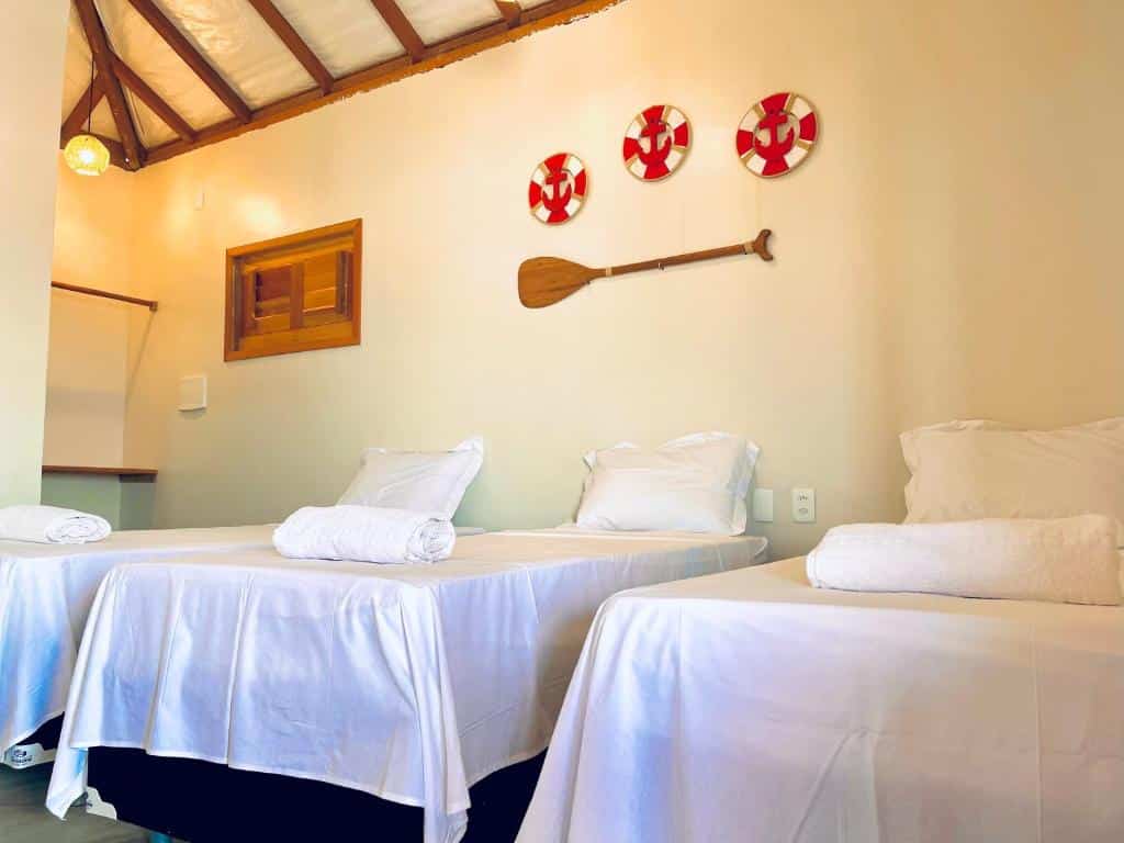 Três camas bem arrumadas com lençóis e toalhas brancos em um quarto da Pousada Ventos do Norte com decoração náutica em uma parede de cor creme. Representa o post de pousadas em Barra Grande no Piauí.