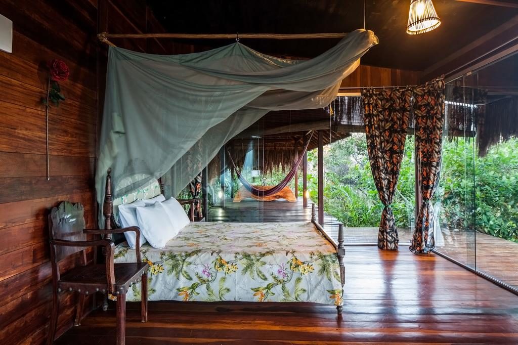 Um quarto de madeira na Pousada O Céu de Boipeba com uma cama de dossel, roupa de cama floral, rede e portas de vidro que se abrem para uma vista da vegetação e uma rede.