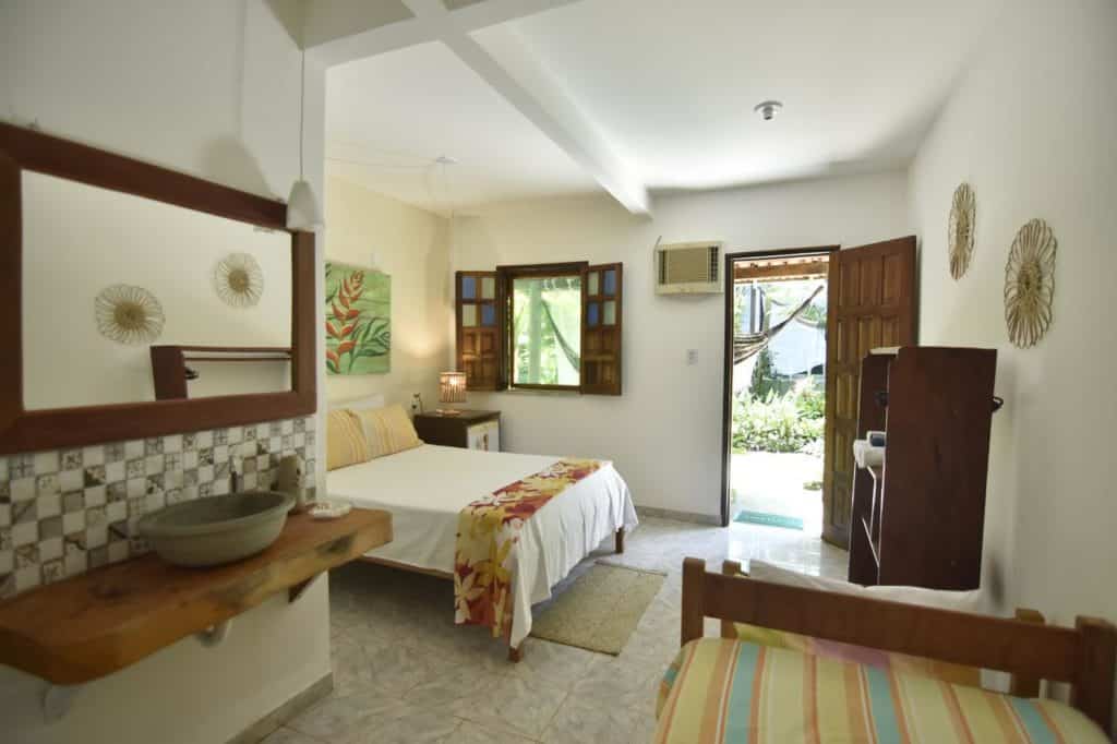 Um quarto arrumado da Pousada Sossego com uma cama de casal, porta e janelas abertas, um espelho de parede e uma pequena mesa de madeira sob a janela.