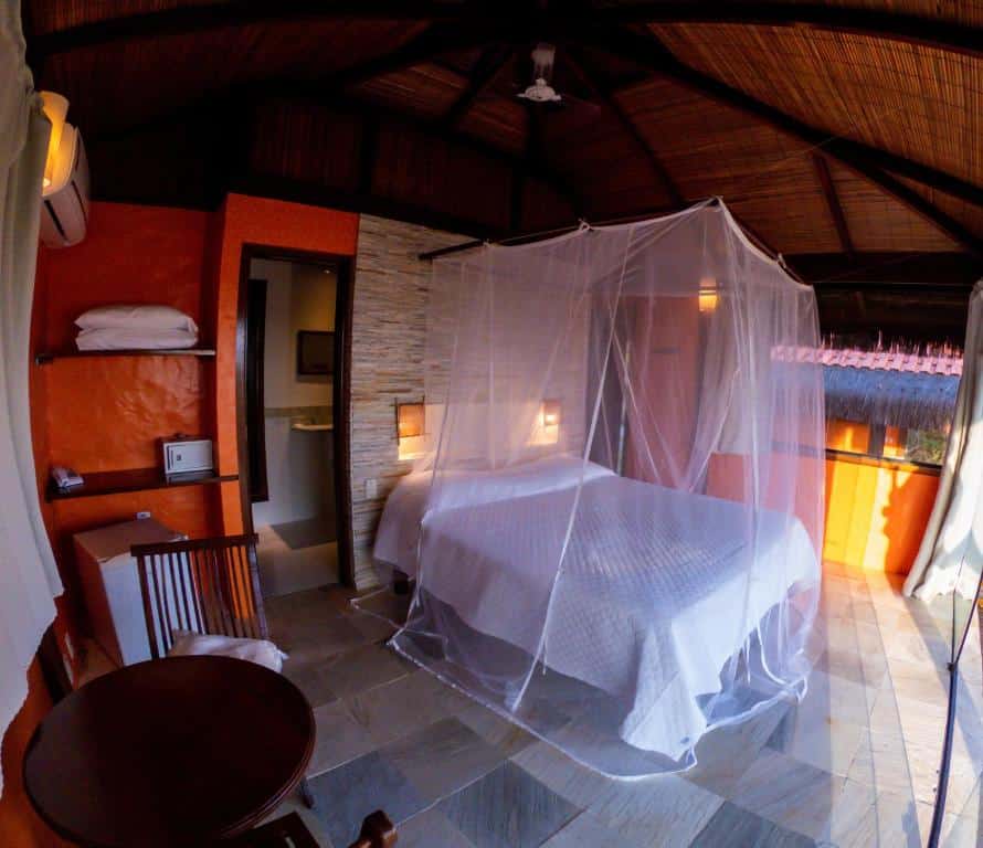 Um quarto da Alizées Moreré com uma cama coberta por um mosquiteiro, uma cadeira de madeira, uma pequena mesa e uma porta aberta para um banheiro.