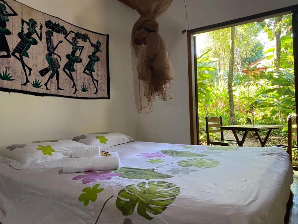 Uma cama na Casa Mestiça com roupa de cama floral, uma toalha em cima e uma pequena mesa com duas cadeiras perto de uma porta aberta que dá para um jardim.
