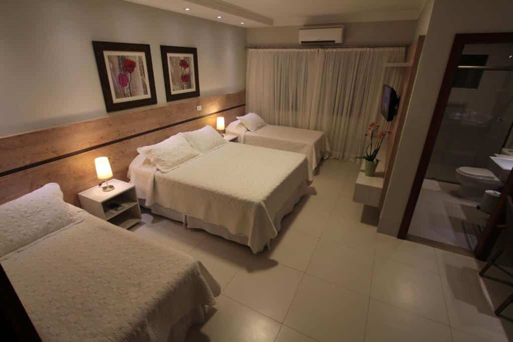 Um quarto da Pousada Surucuá com três camas, mesinhas de cabeceira com luminárias, arte emoldurada e um banheiro privativo visível à direita. Esta é uma das pousadas em Bonito.