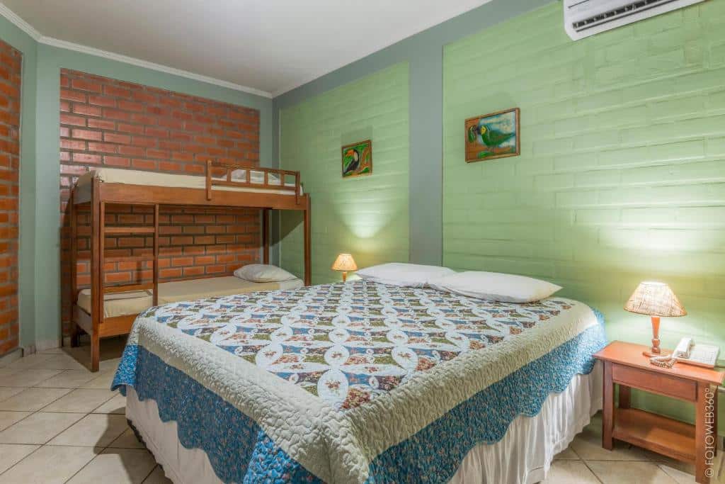 Quarto da Pousada Rancho Jarinu com uma cama de casal, beliches, duas mesas laterais com luminárias e duas pinturas em paredes verdes e de tijolos. Esta é uma das pousadas em Bonito.