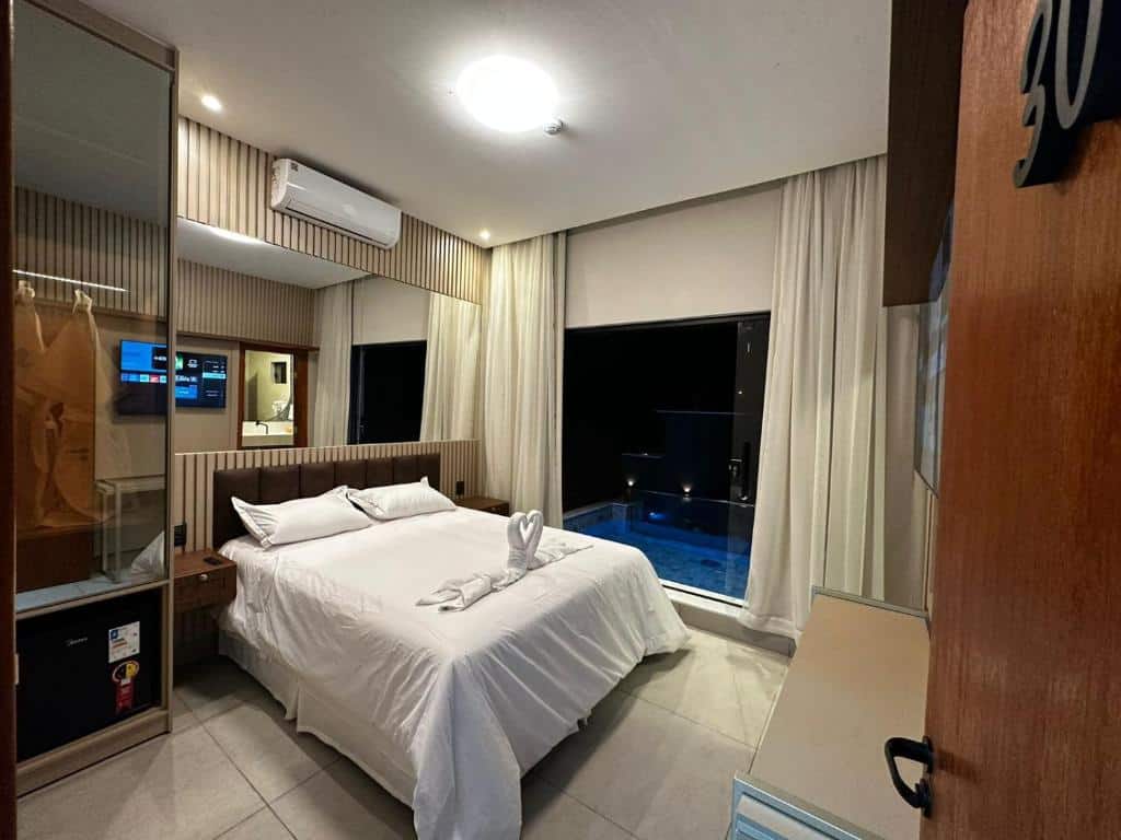 A imagem mostra o interior de um quarto da Pousada Villas em Bonito, Mato Grosso do Sul. O quarto é decorado de maneira moderna e aconchegante, com tons neutros e detalhes em madeira. A cama de casal é coberta com roupas de cama brancas impecáveis e há uma toalha dobrada em forma de cisne sobre a cama. À esquerda, há um armário com portas de vidro espelhado e um frigobar embutido. Acima do frigobar, há um suporte para malas. Na parede oposta à cama, há uma televisão de tela plana montada, permitindo entretenimento para os hóspedes. O quarto possui ar-condicionado montado na parede para maior conforto. Uma grande janela com cortinas claras oferece uma vista para a área externa, onde se pode ver uma piscina privativa iluminada à noite, proporcionando um ambiente relaxante e luxuoso.