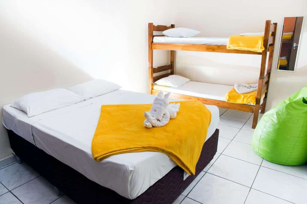 Quarto da Pousada São Jorge com uma cama de casal, beliche, cobertores amarelos, um pufe verde e piso de azulejos brancos.