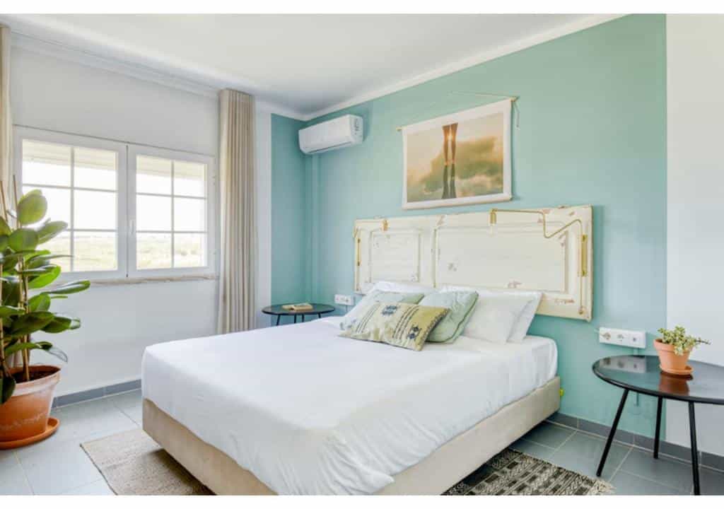 Quarto da da Selina Bonito com uma cama de casal, parede com detalhes verdes, vasos de plantas, janela e um ar-condicionado acima da cama.