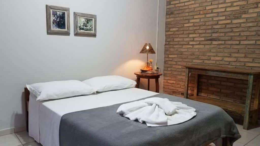quarto da Pousada Cheiro de Mato com uma cama com roupa de cama branca e cobertor cinza, uma toalha dobrada em cima, uma mesa de cabeceira com uma lâmpada e uma parede de tijolos.