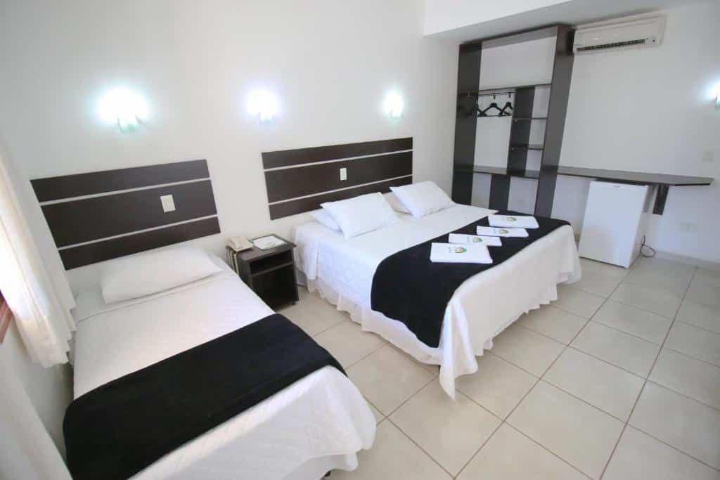 Quarto da Pousada Remanso com uma cama de solteiro e uma cama de casal, roupa de cama branca, detalhes em preto, frigobar pequeno e armário aberto.