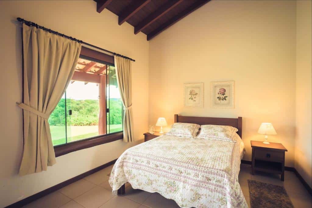 Um pequeno quarto da Pousada Boyrá com uma cama de casal, roupa de cama floral, duas mesinhas de cabeceira com luminárias e uma janela com vegetação externa.