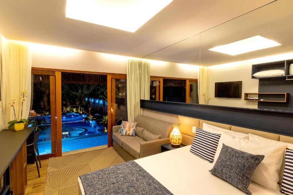 Quarto da Pousada Arte da Natureza com cama, sofá, TV e janelas grandes com vista para a área da piscina iluminada à noite.