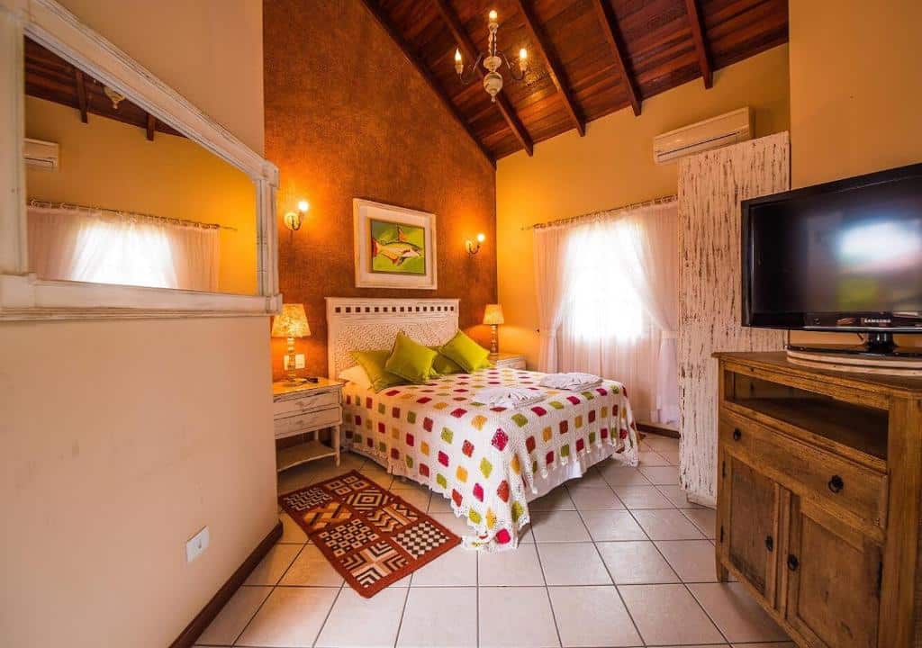 Quarto aconchegante da Pousada Águas de Bonito com uma cama de casal, colcha estampada, TV em uma cômoda de madeira e iluminação aconchegante sob um teto abobadado.