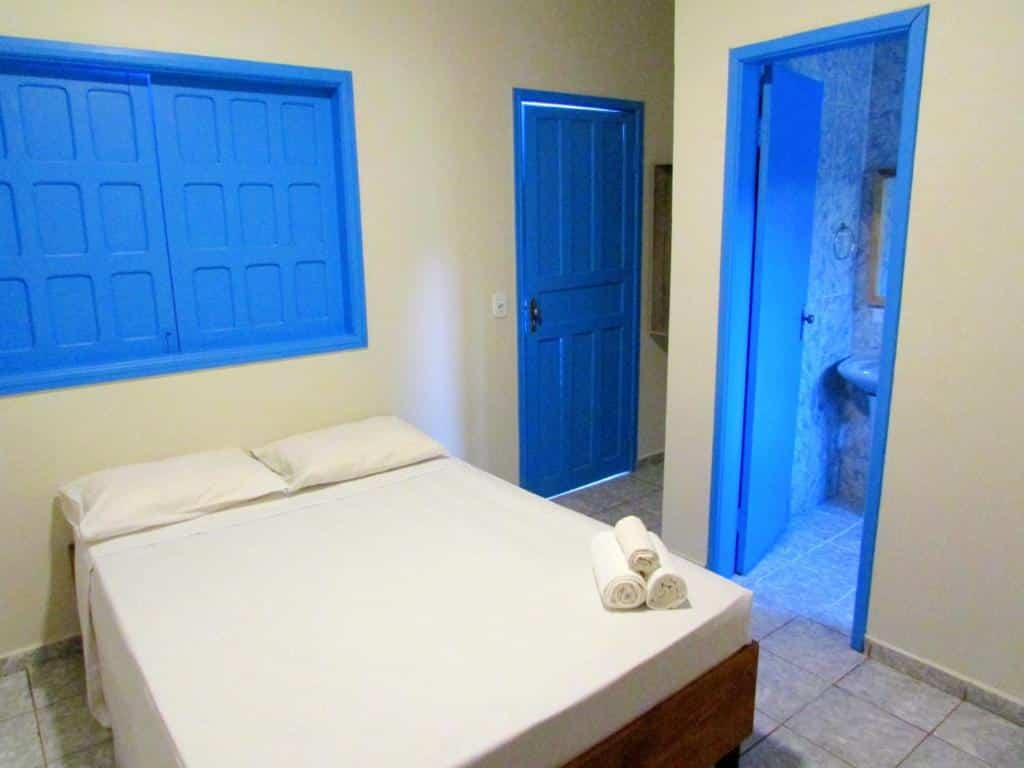 Um quarto da Pousada Cambará com uma cama de casal, roupa de cama branca, portas e janelas azuis e uma porta aberta para um banheiro de azulejos.