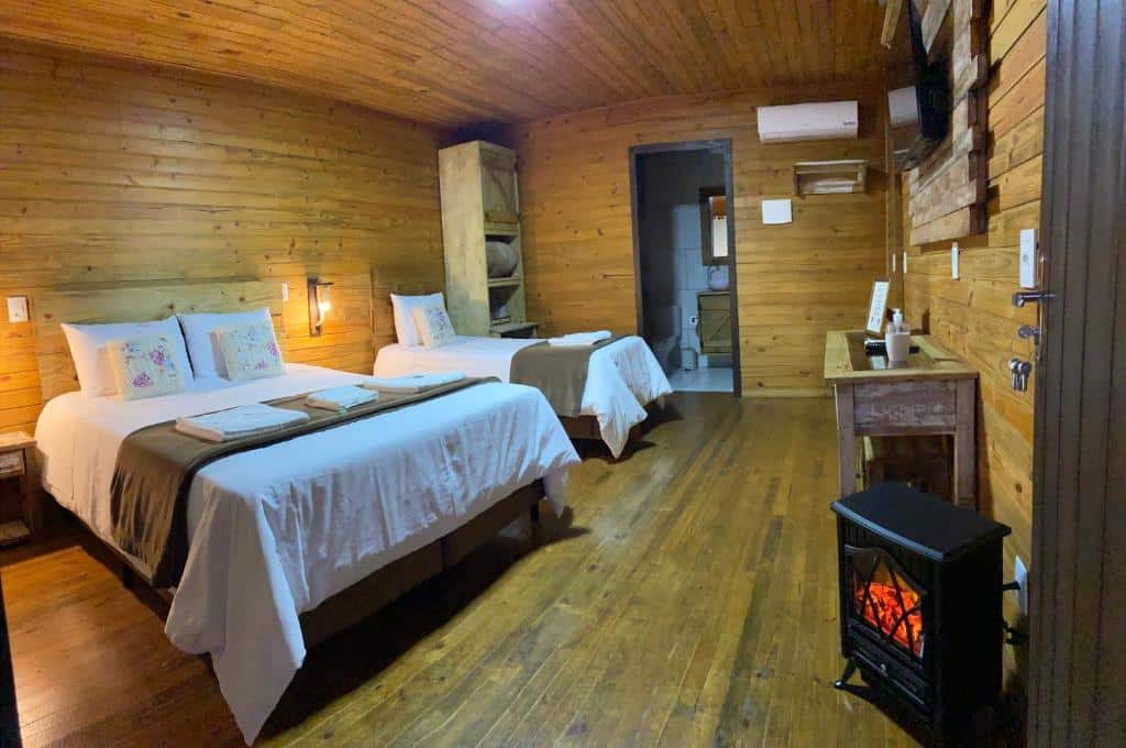 interior do chalé na Pousada Costaneira, uma das pousadas em Cambará do Sul, com uma cama de casal, uma cama de solteiro, uma lareira móvel pequena à direita e uma porta com vista para o banheiro na parede dos fundos do quarto