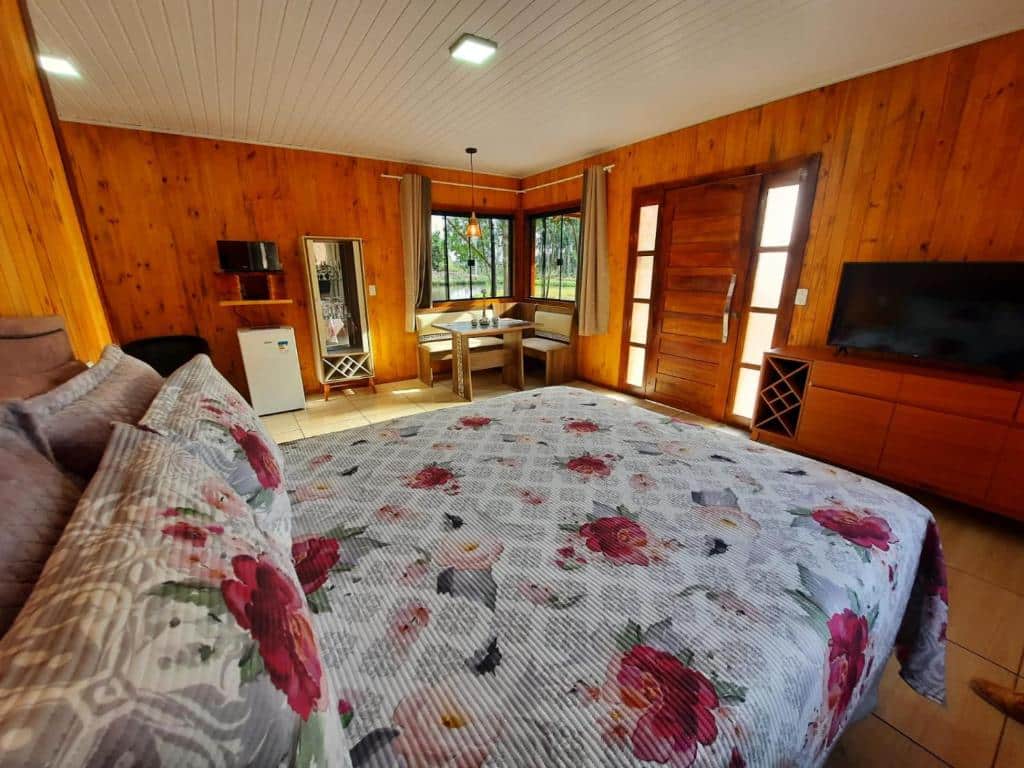 vista interna do quarto do Cânions de Cambará com uma cama de casal, mesa e cadeiras de canto com janelas amplas, frigobar e televisão de tela plana